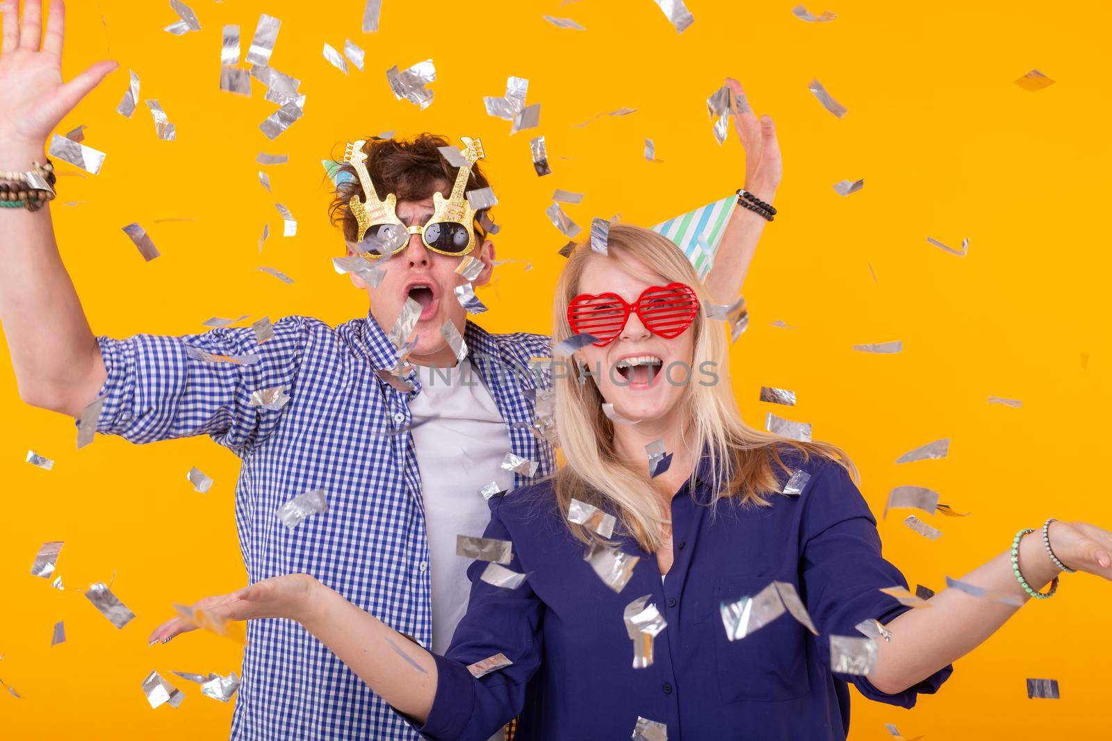 Young cheerful positive couple of funny glasses and a paper cap rejoice and shout on a yellow background with flying confetti. Conception of a holiday and fun