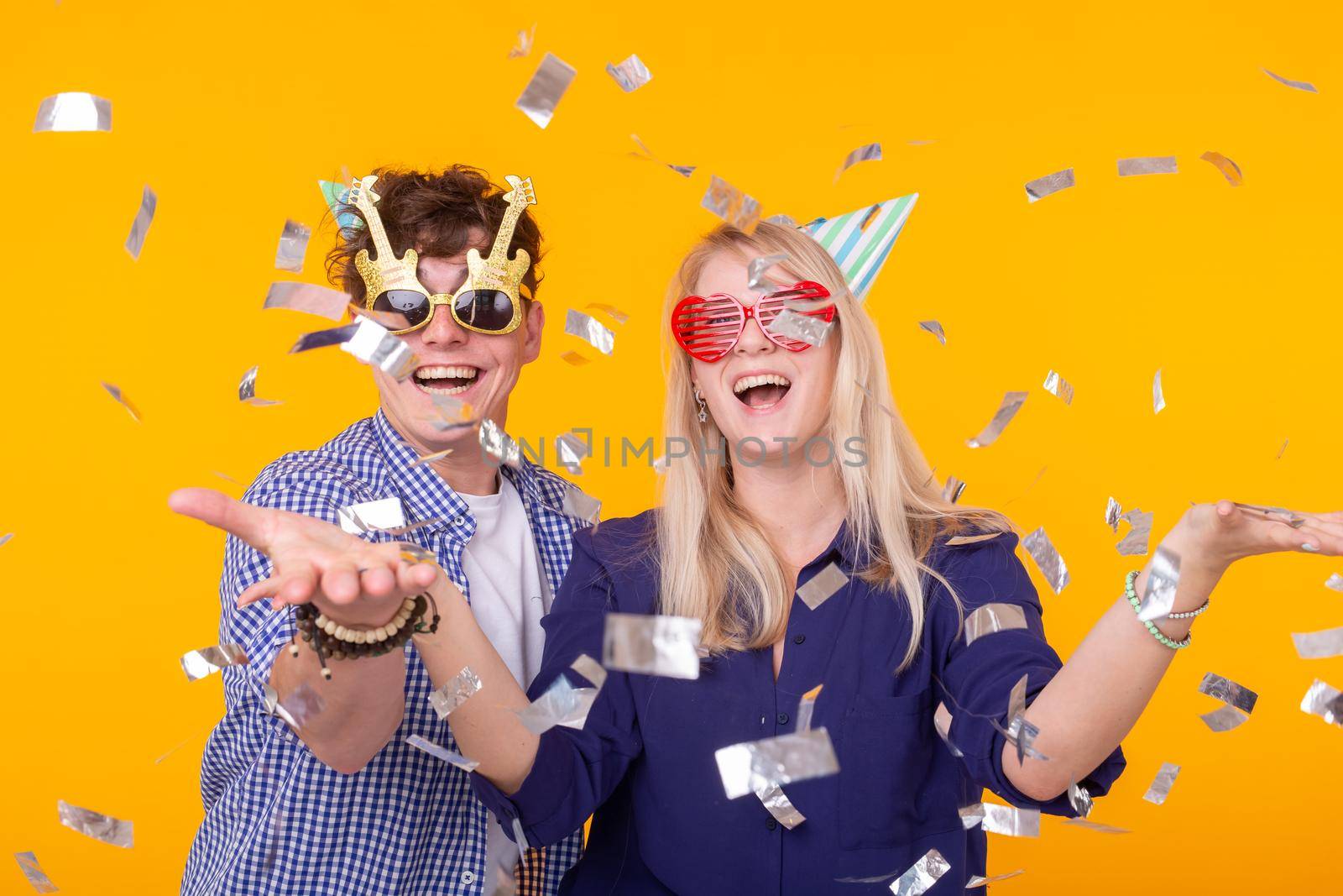 Young cheerful positive couple of funny glasses and a paper cap rejoice and shout on a yellow background with flying confetti. Conception of a holiday and fun