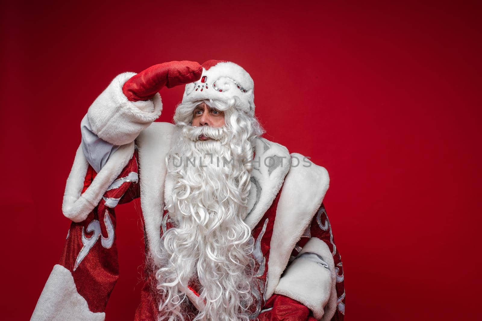 Festive Santa Claus look into the distance with hand on forehead on red background, copy space for holiday advertising by StudioLucky