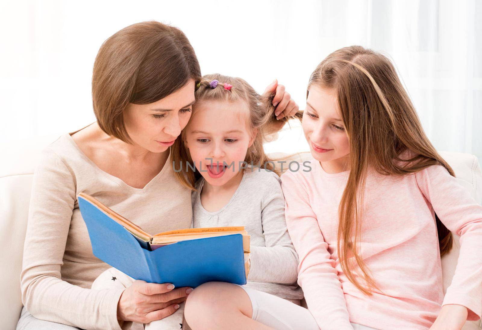 Mom and her two little daughters reading children's book in the living room. Mom and daughters sitting on the coach and reaading