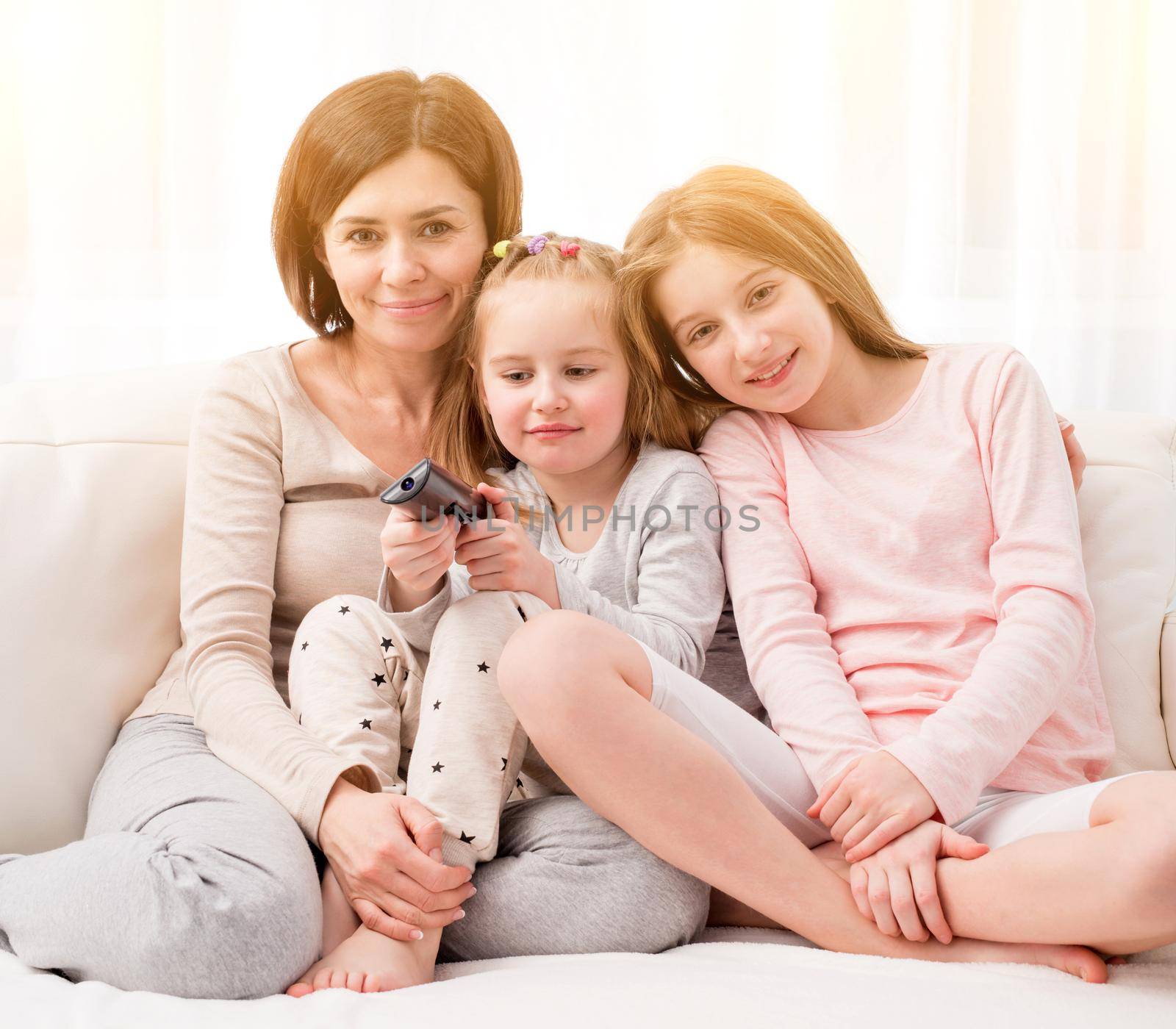 Portrait of mom hugging her cute daughters while sitting on the sofa and watching TV at home. Happy family relaxing on the coach