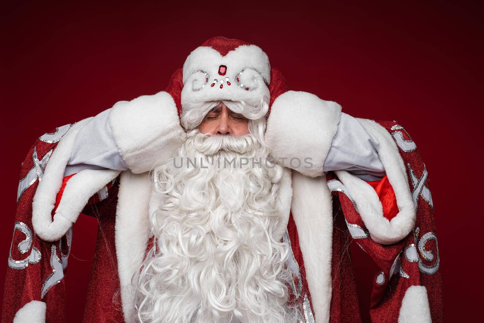 Santa Claus get tired and grabbed head for a headache, closed eyes, studio portrait on red background by StudioLucky