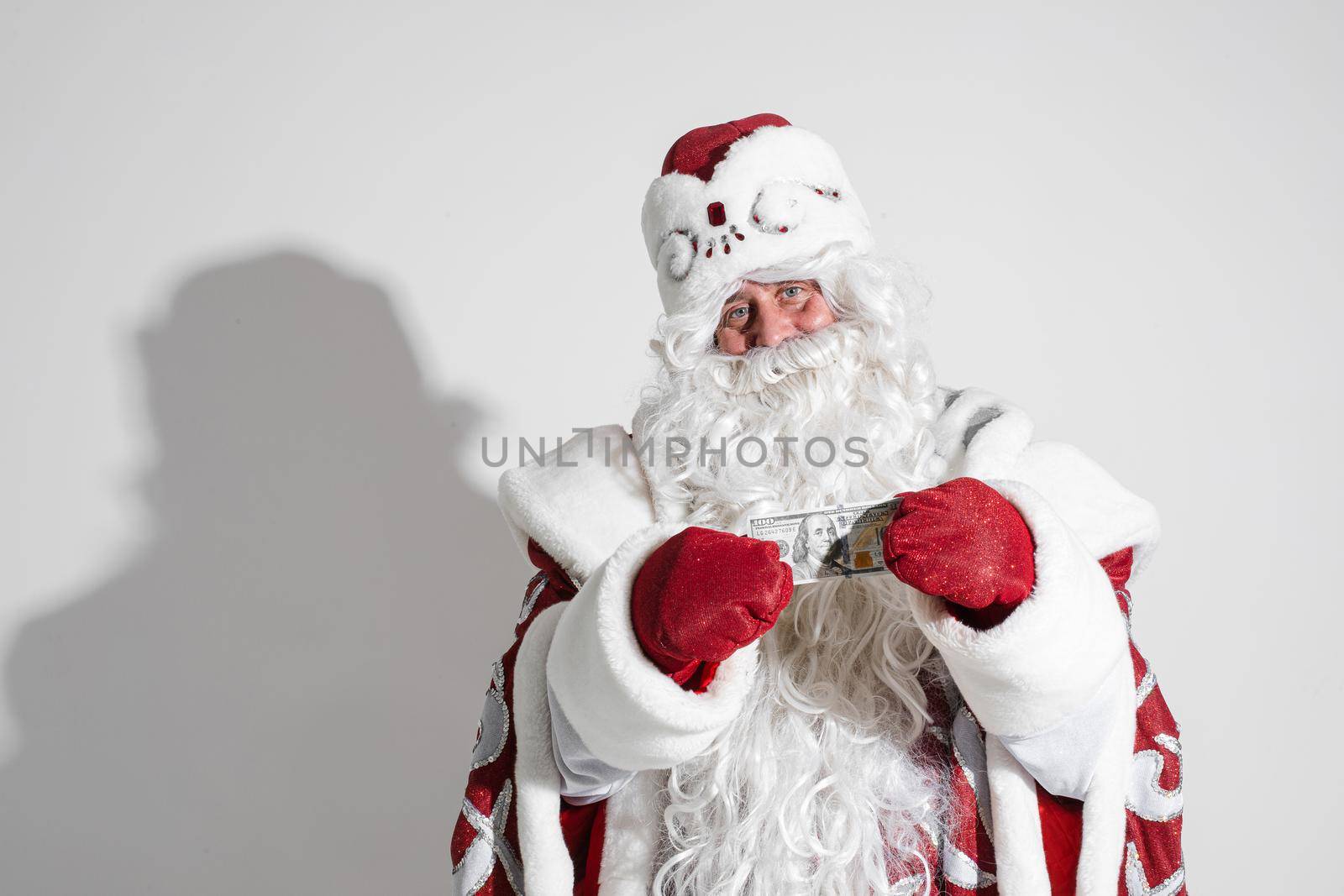 Smiling generous Santa Claus with long white beard in fancy costume holding banknote in hands wearing red mittens. Cutout on white background.