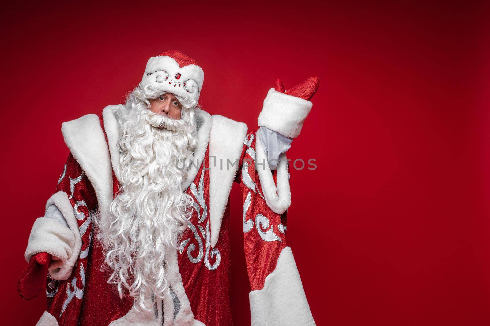 Santa Claus elderly male in festive xmas costume point by hand to copy space on red background by StudioLucky