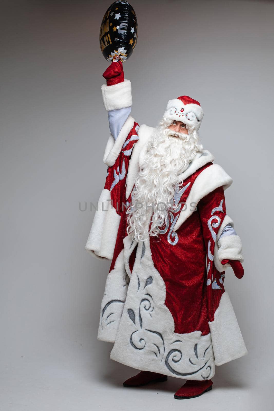 Stock photo of a Father Frost with long white beard in traditional red costume with hat and mittens holding inflatable air balloon in right hand raised up.