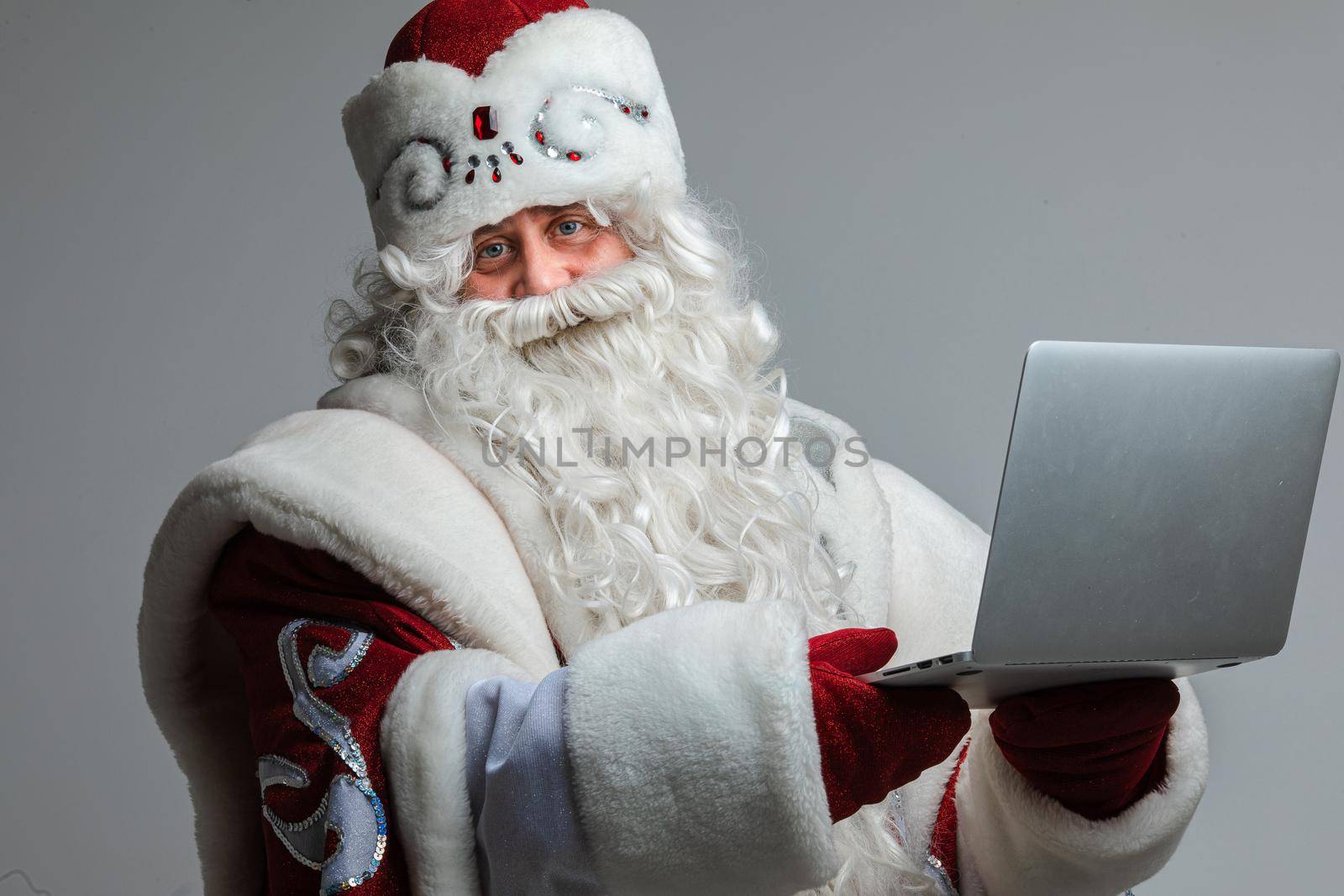 Stock photo of smiling happy Father Frost with white long beard in festive winter hat and traditional coat holding laptop and looking at camera.