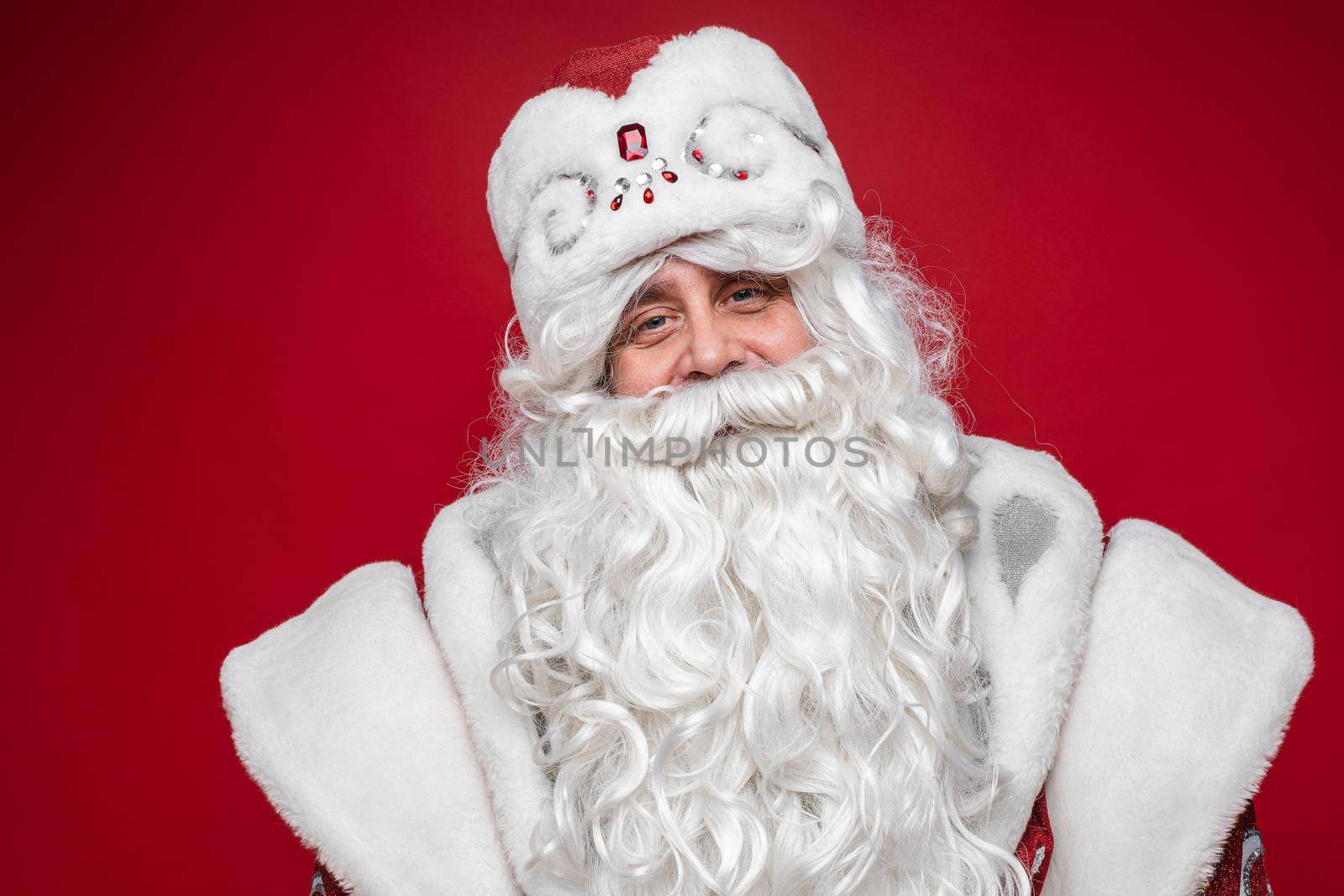 Stock photo of smiling happy Santa in traditional hat with rhinestones and long white beard looking at camera. Cutout on red background. Happiness concept.