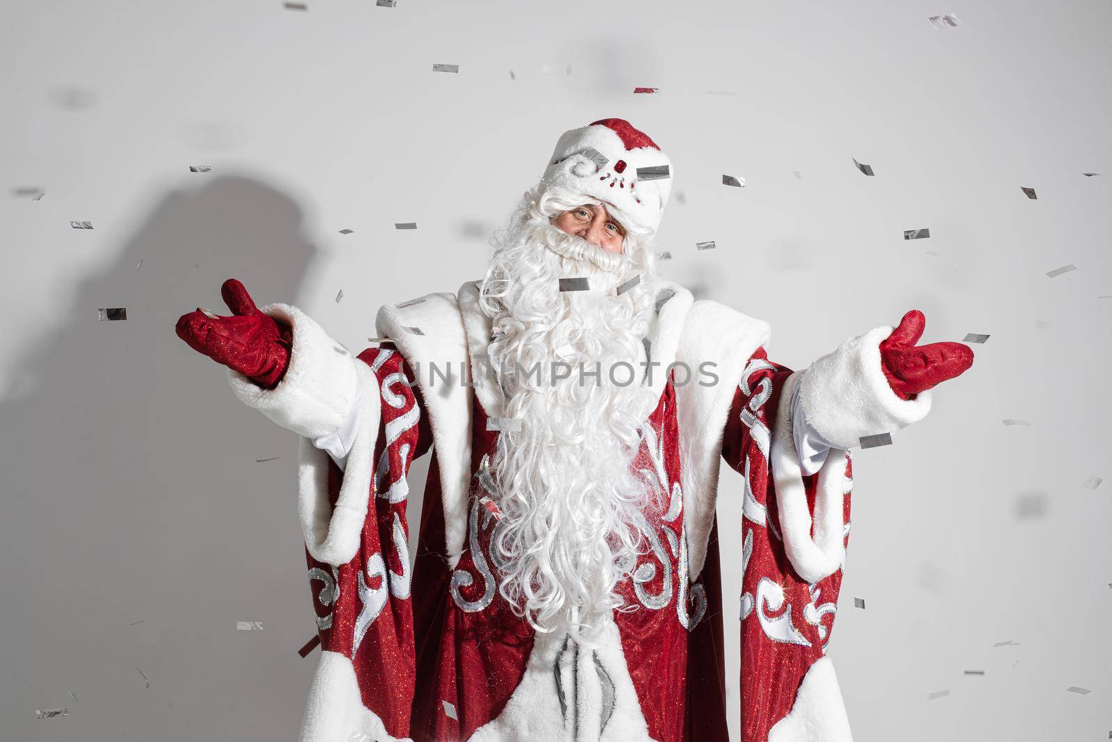 Santa in red traditional costume and white beard outstretching arms under falling silver confetti. Father Christmas congratulating with New Year.