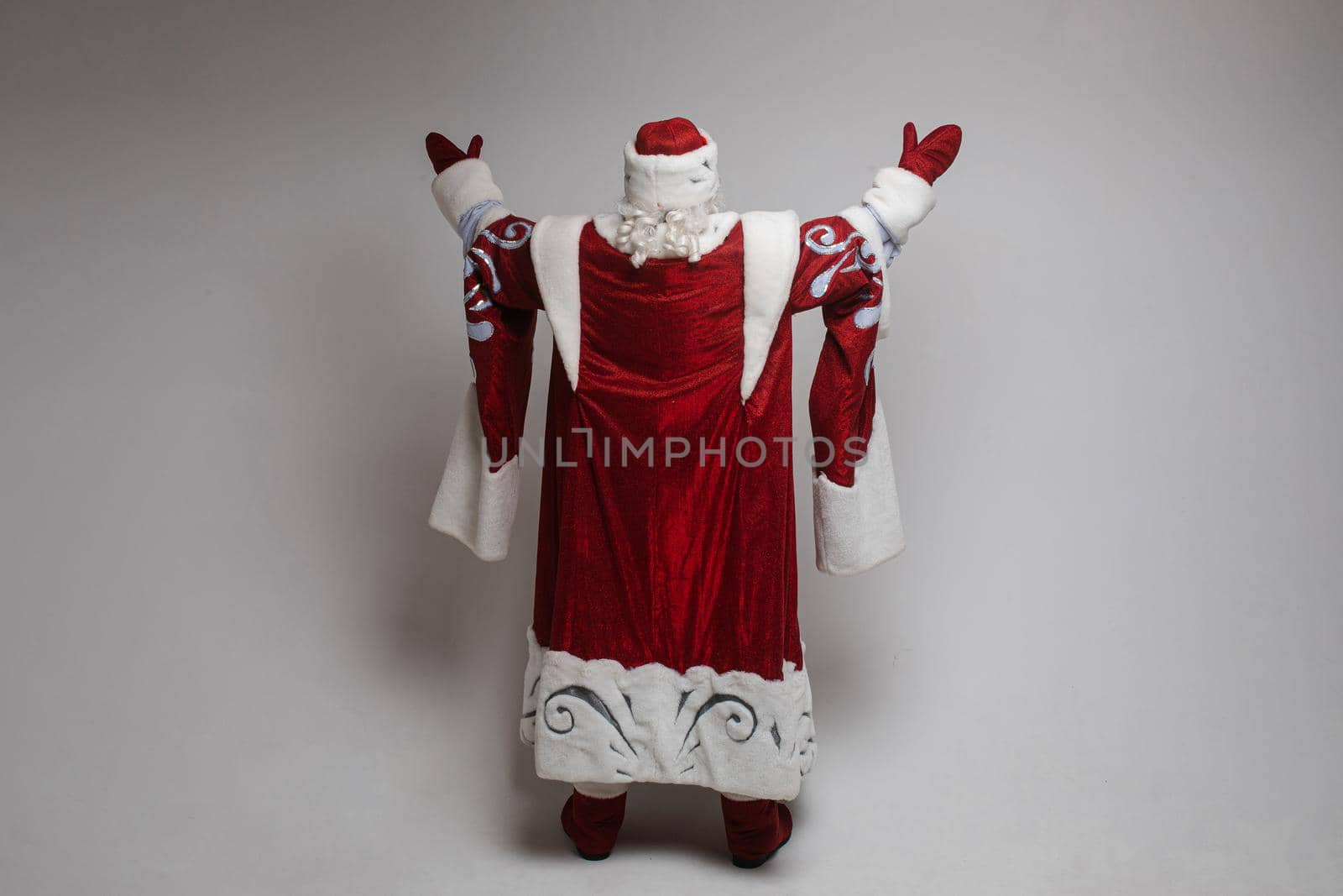 Studio photo of unrecognizable Father Frost in traditional red and white costume holding arms up against grey background.