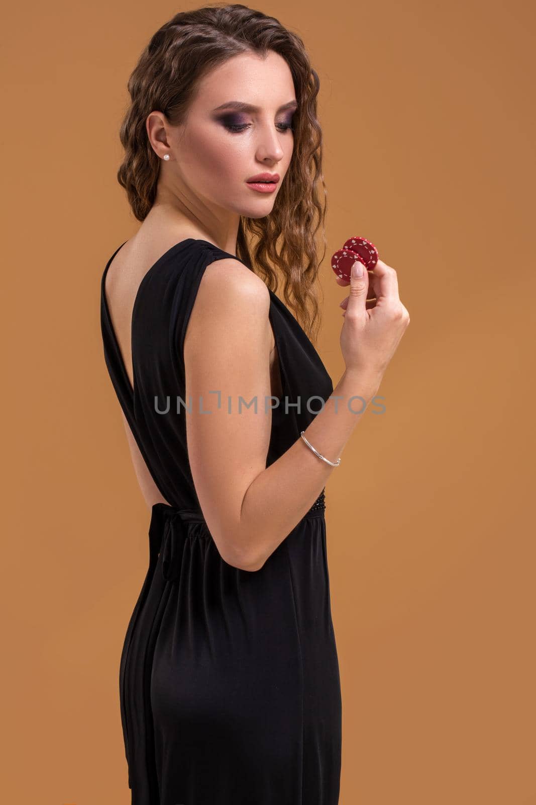 Pretty long hair woman in black dress holding chips for gambling on beige background. Studio shot