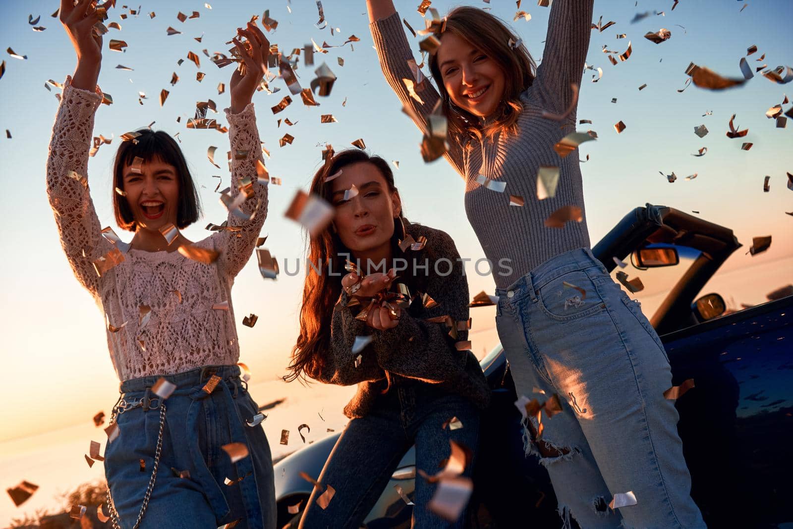 Cheerful young three women are dancing near the cabriolet. They throw up falling leaves. Beautiful sunset at the background