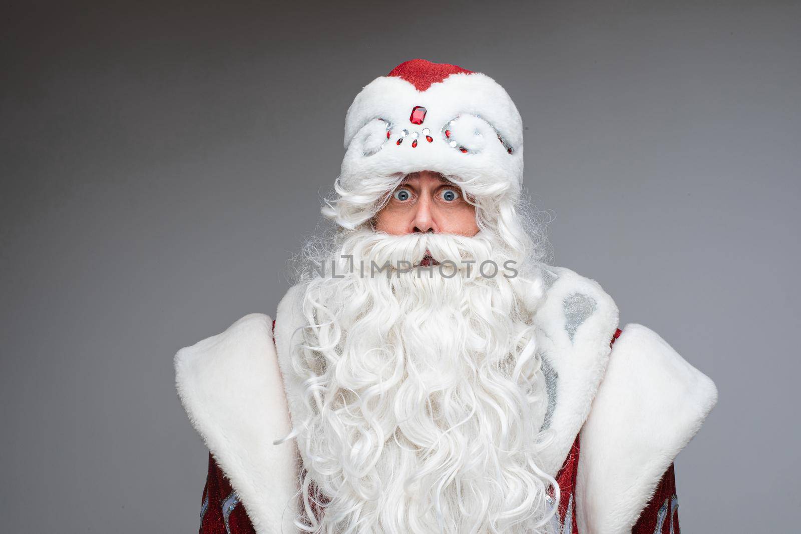 Portrait of worried Santa Claus in fancy costume with white long beard looking at camera with eyes wide open. Cutout on grey background.