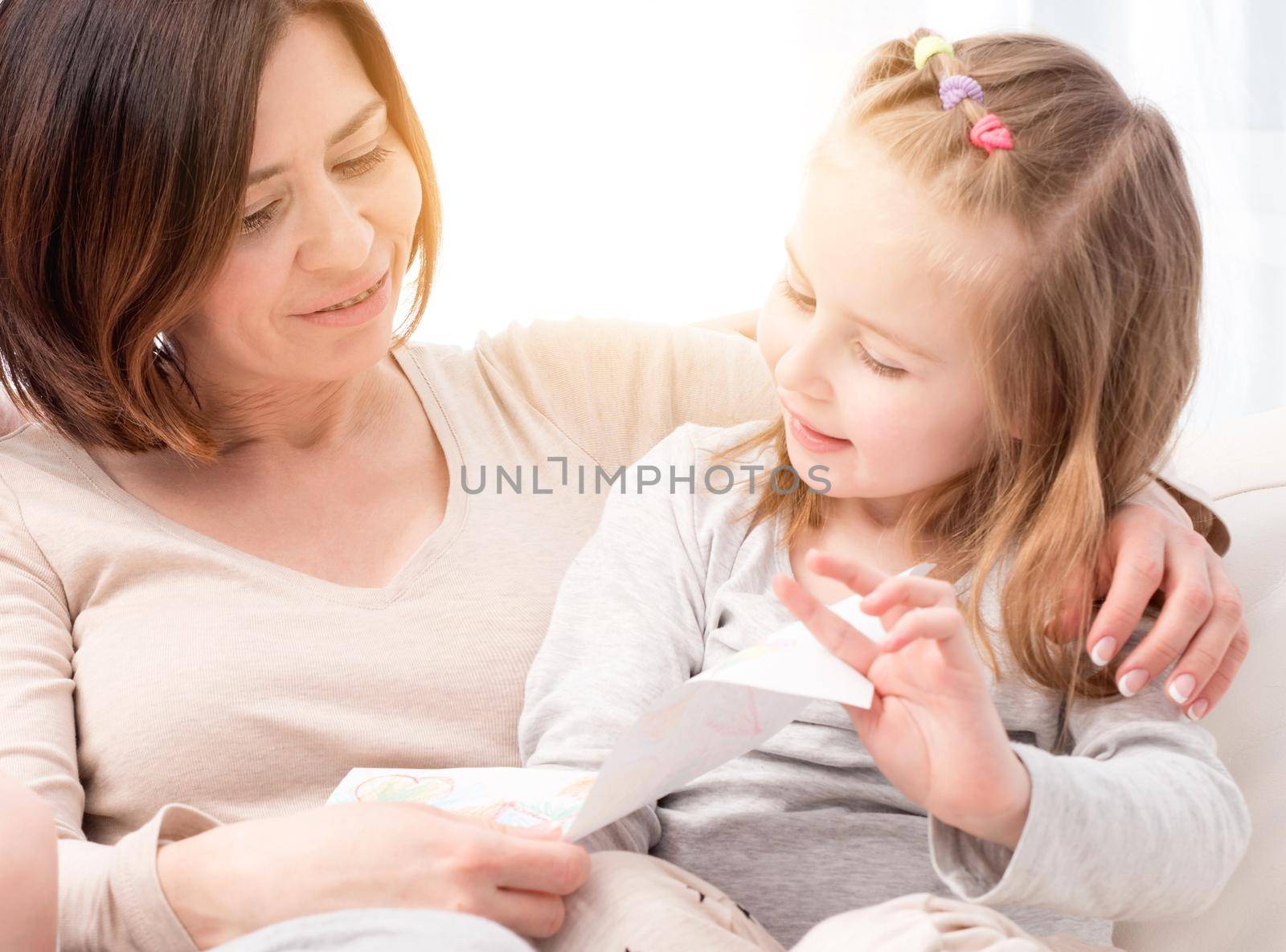 Happy smiling mom looking at the mother's day greeting card made by daughter sitting on sofa with little child