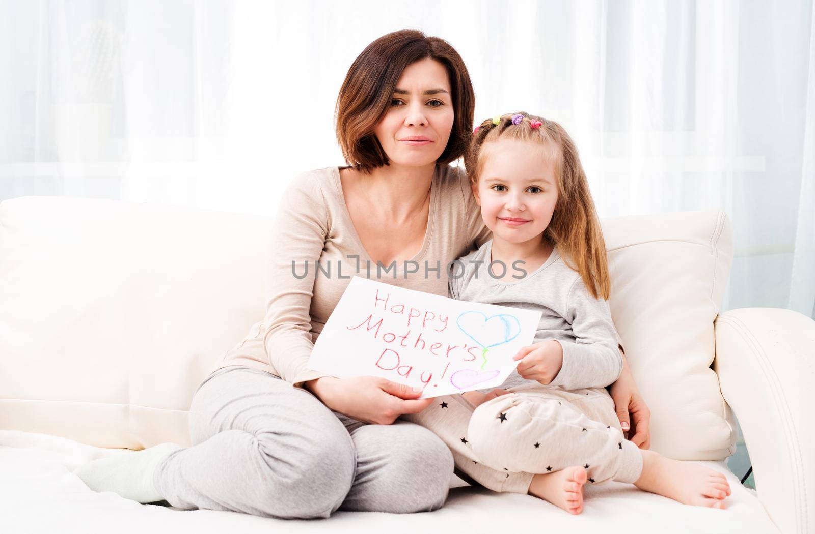 Happy Mother's Day! Mom and daughter sitting on the sofa and holding hand-made drawing made by little daughter