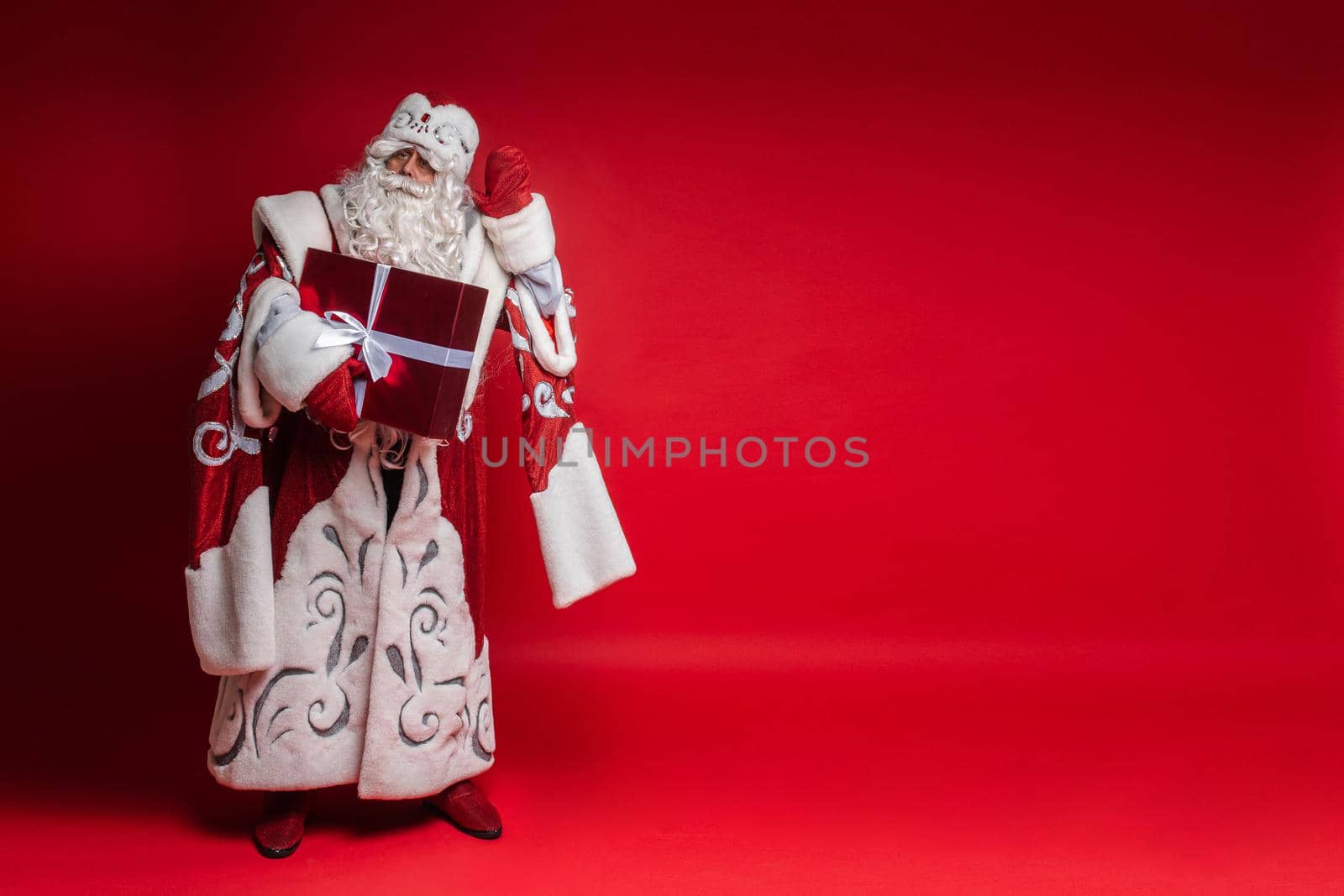 Santa Claus with xmas gift puts his hand to head, tryes to listen on red background with copy space for festive design. High quality photo