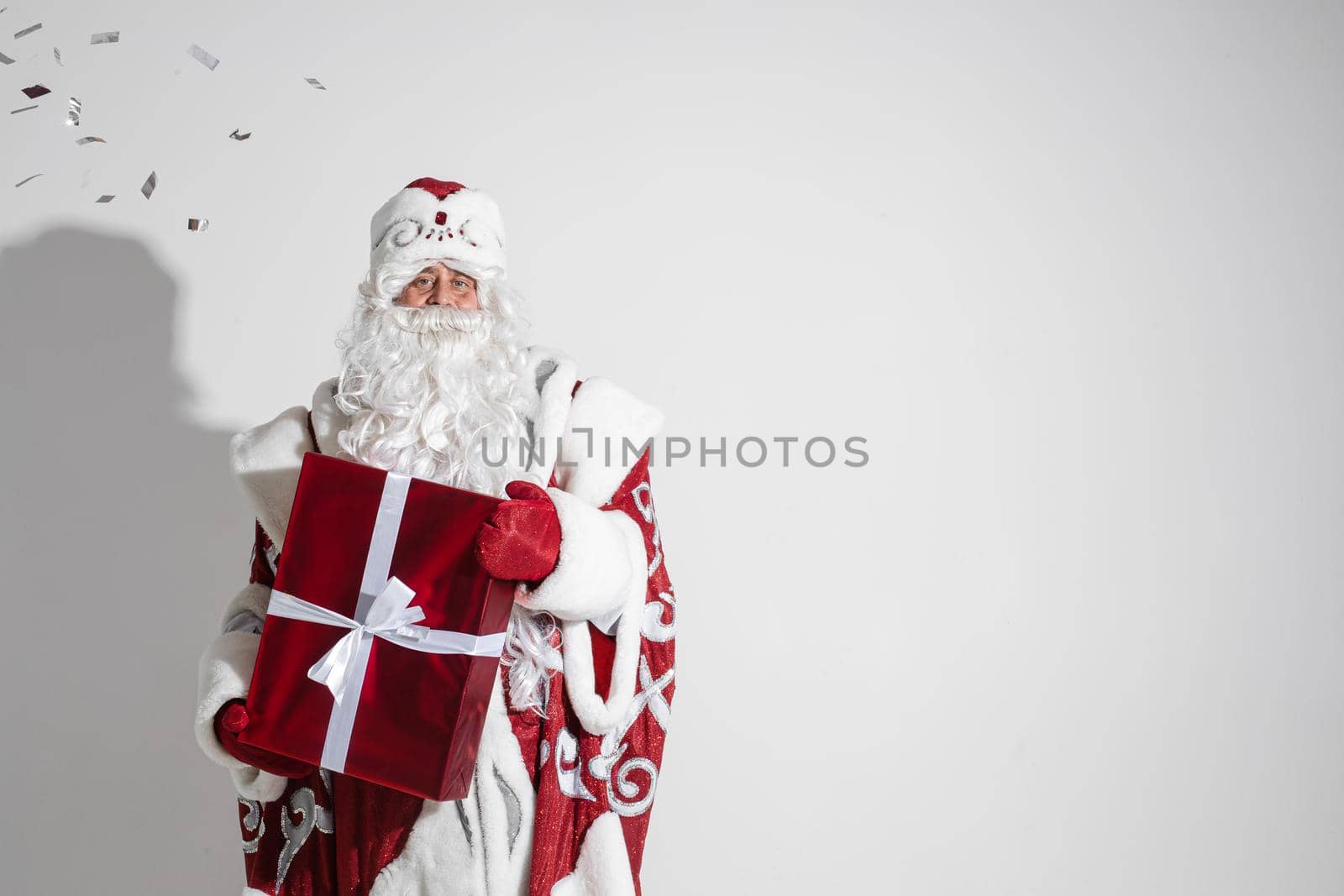 Santa Claus with red xmas gift posing in studio, elderly male with christmas present, copy space for holiday advertising. High quality photo