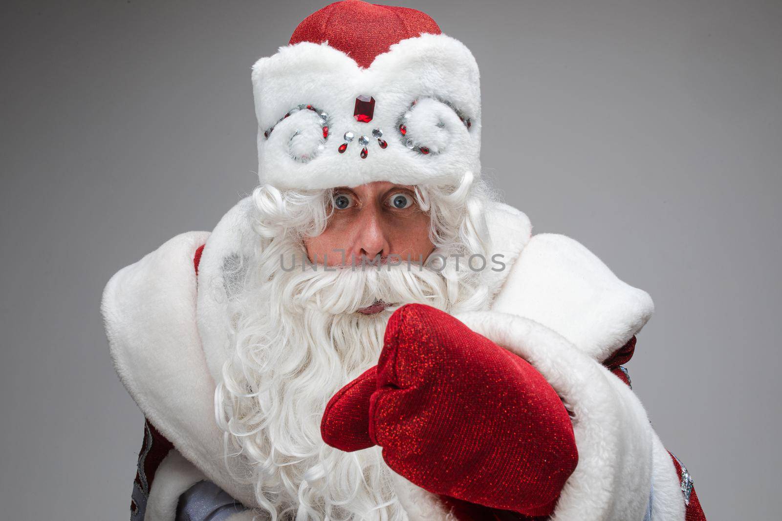 Portrait of Santa Claus with white long beard in fancy costume pointing at camera with red mitten on his hand looking surprised or shocked.