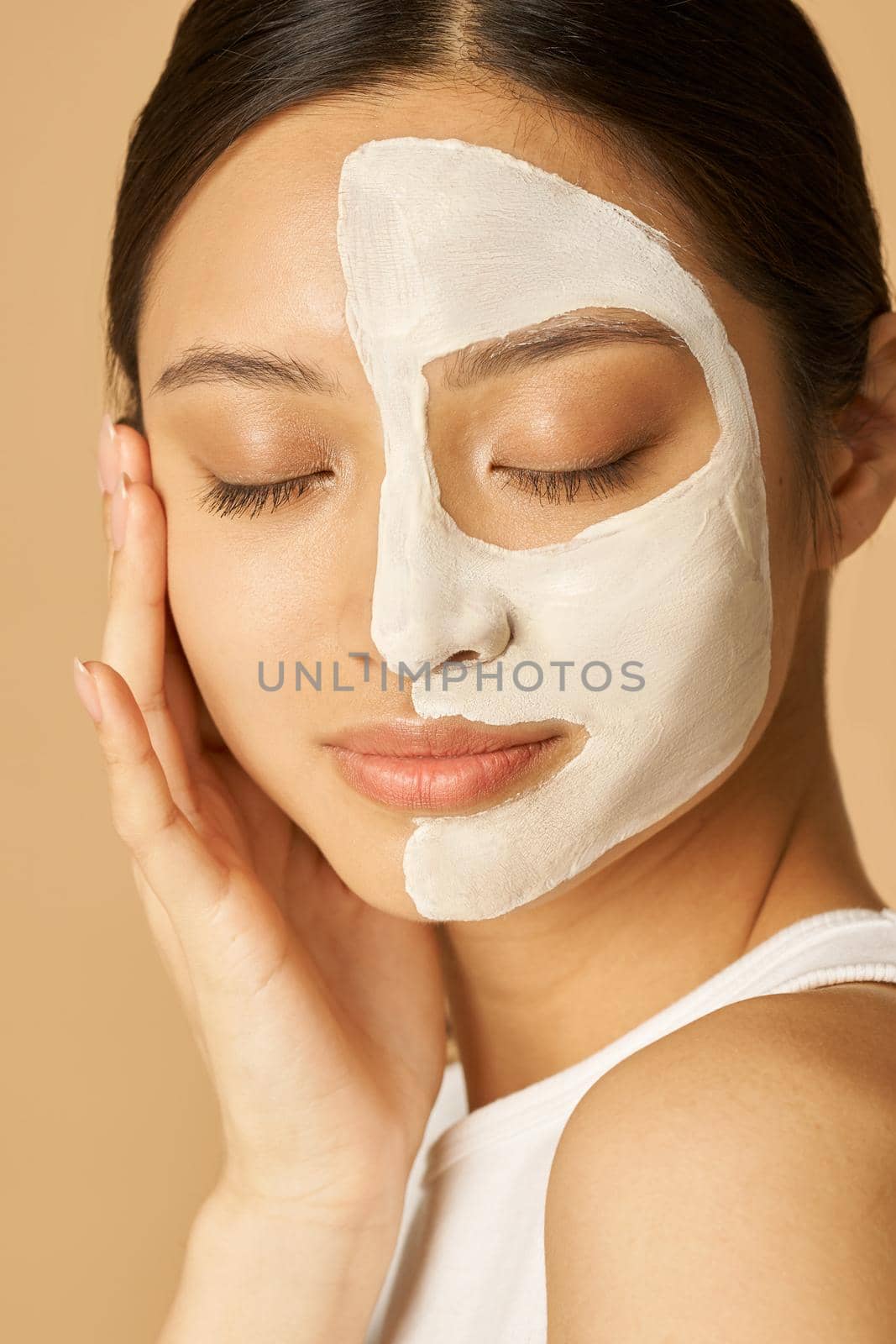 Relaxed young woman with facial mask applied on half of her face receiving spa treatments, posing for camera with eyes closed isolated over beige background by friendsstock
