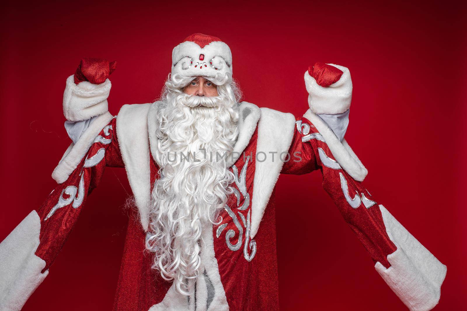 Serious senior male in xmas Santa Claus costume posing on red background celebrating new year by StudioLucky