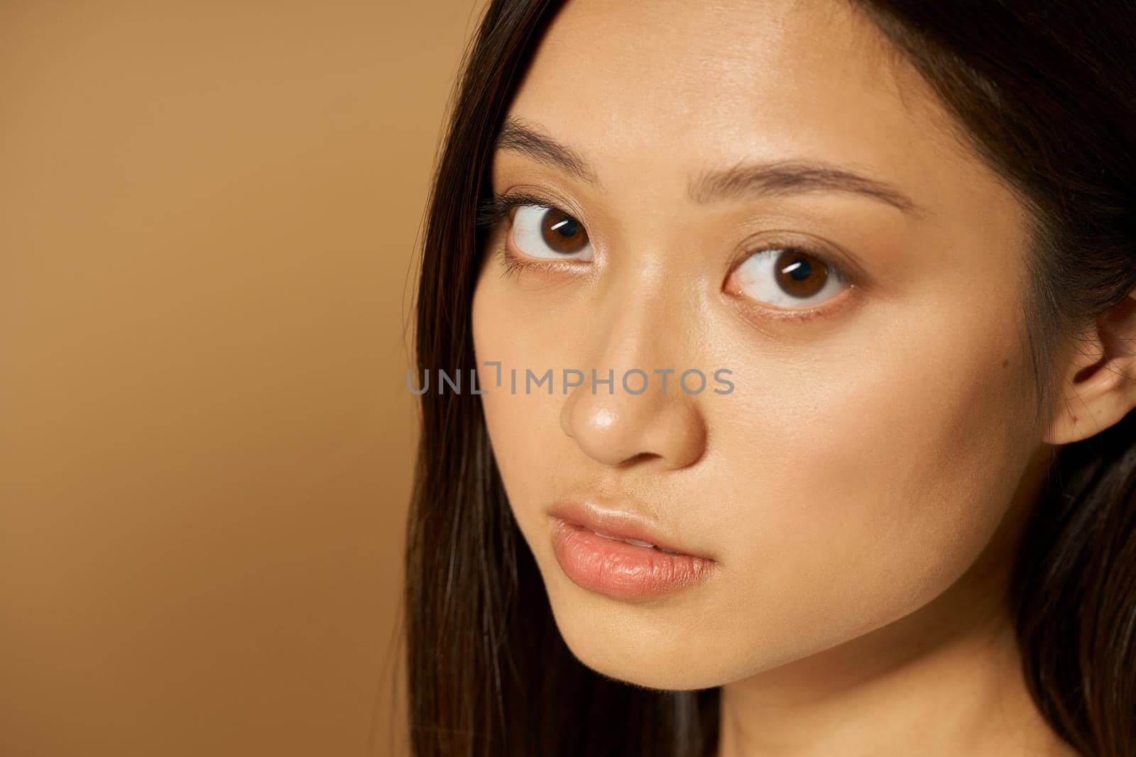 Face closeup of beautiful mixed race young woman with glowing skin looking at camera while posing isolated over light brown background. Natural beauty, skincare concept