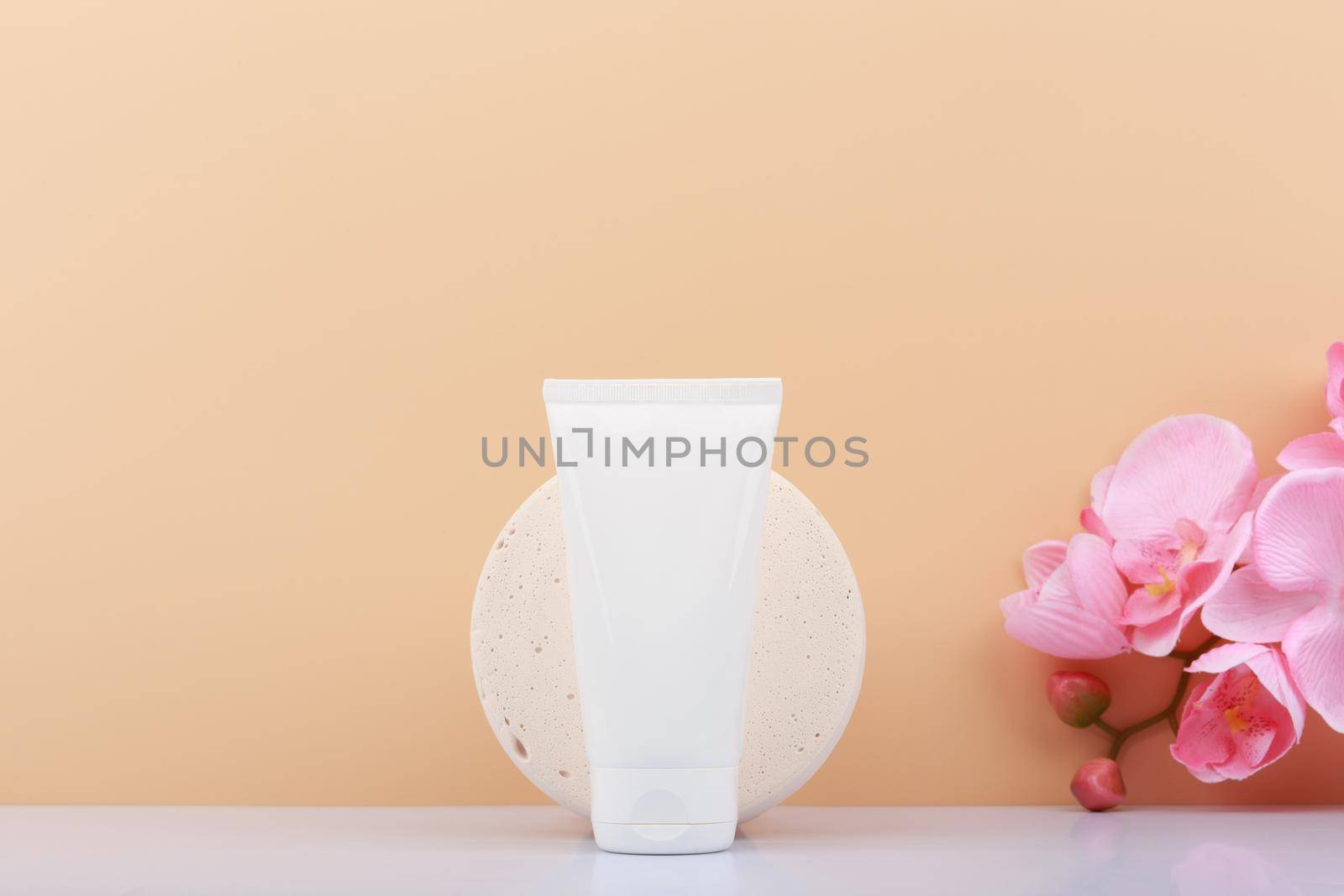White plastic cosmetic tube with cream, balm, mask, scrub or lotion on beige background decorated with pink flower with copy space. Concept of natural organic skin care products for manicure, hands or face