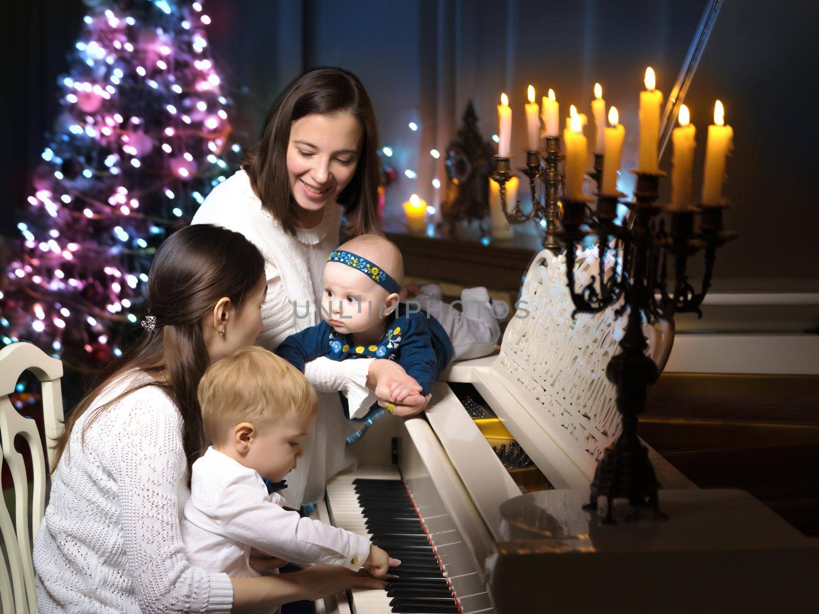 Young mothers with their young children in a dark room by candlelight, near a large white grand piano. The concept of holidays and happy childhood.