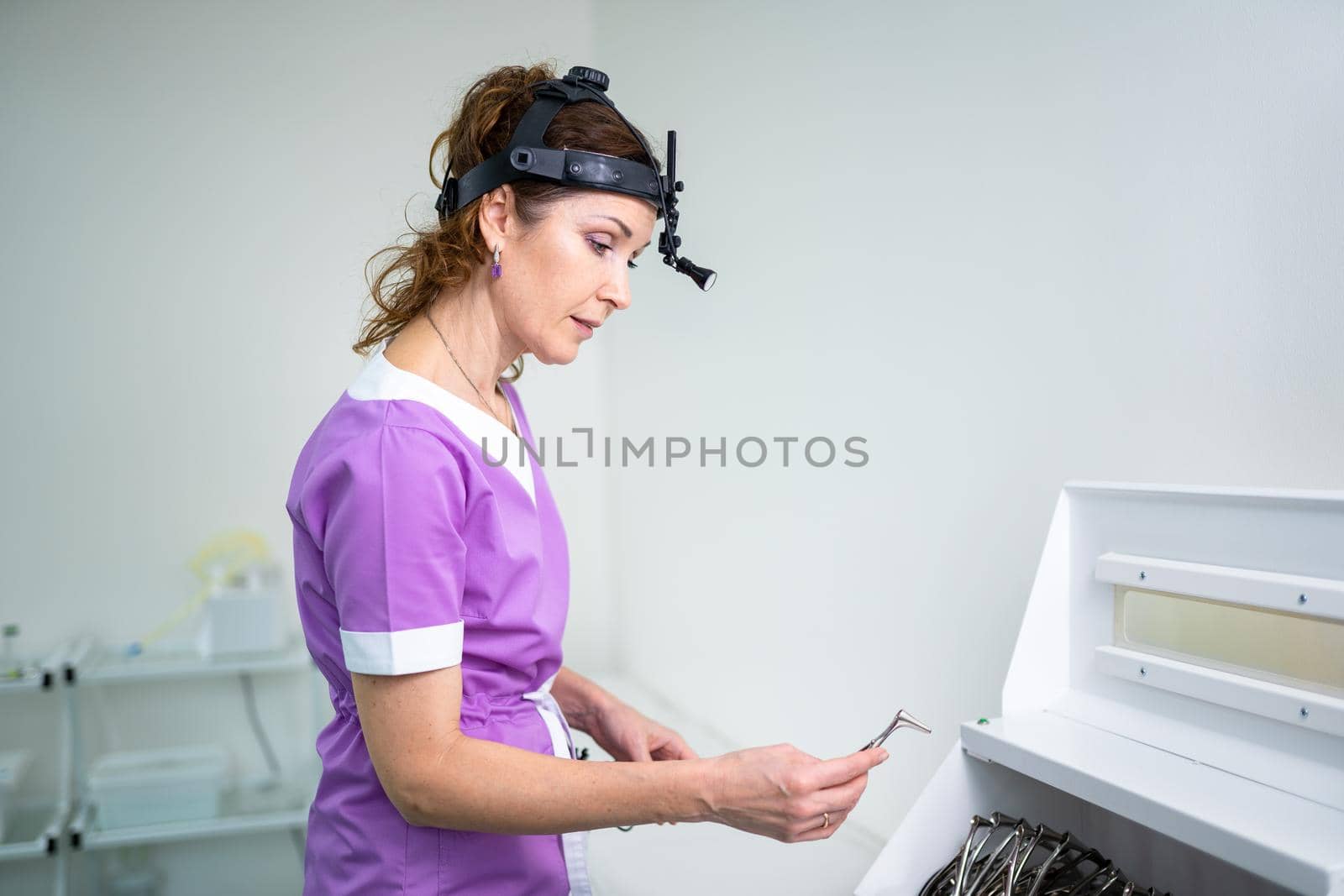 Portrait of confident female doctor. Female ENT doctor in purple medical clothes posing in office of the clinic on her head with a sedecin instrument to examine the patient. Healthcare occupations by Tomashevska