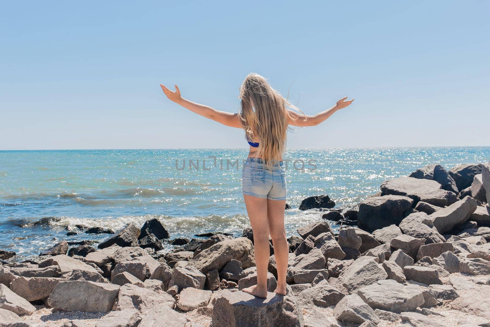 A young teenage girl with long blonde hair blonde in short shorts raised and spread her hands to the sides standing on the rocks near the sea shore and the beach. The concept of freedom by AYDO8