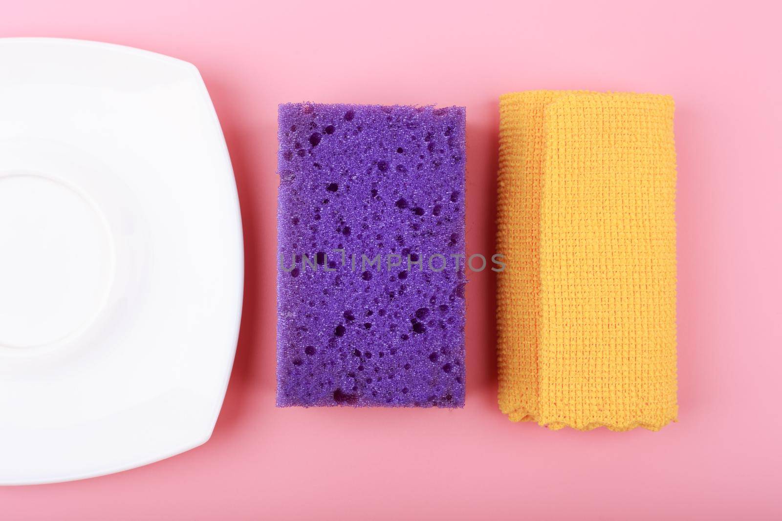 Creative flat lay with yellow dust cloth, white plate and purple cleaning sponge on pink background. House cleaning and dishwashing concept