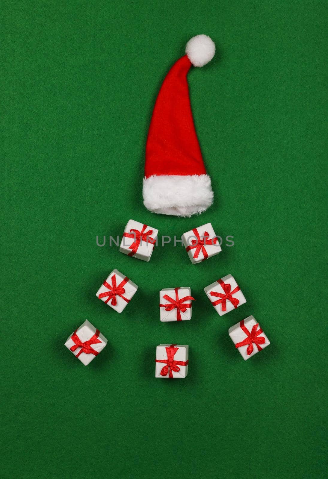 Close up white giftboxes Christmas decoration out of red Santa hat over green felt background with copy space, table top view, flat lay