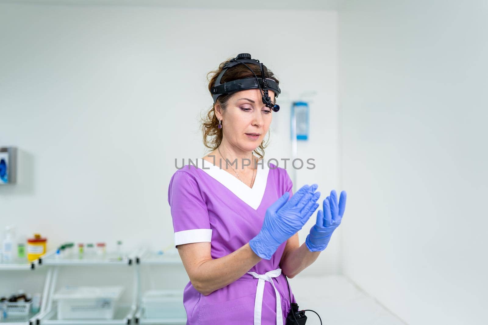 Medical worker of ENT clinic middle aged Caucasian female in purple medical uniform posing looking at the camera in the examination room. Professional medical specialist otolaryngologist in hospital by Tomashevska