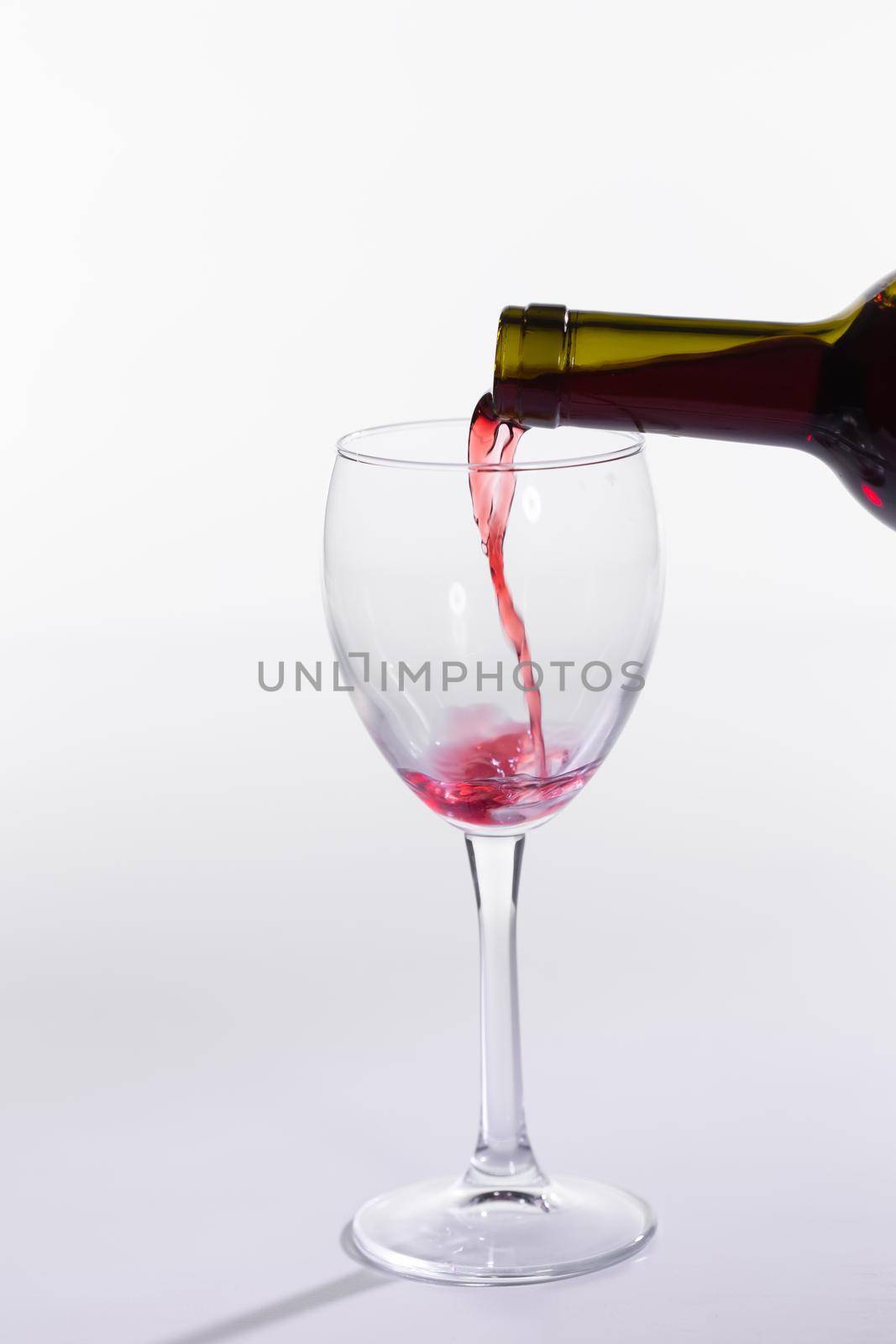 Red wine pouring from bottle into big glass on white background.