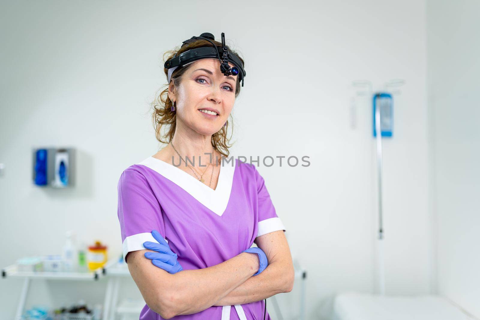 Medical worker of ENT clinic middle aged Caucasian female in purple medical uniform posing looking at the camera in the examination room. Professional medical specialist otolaryngologist in hospital.