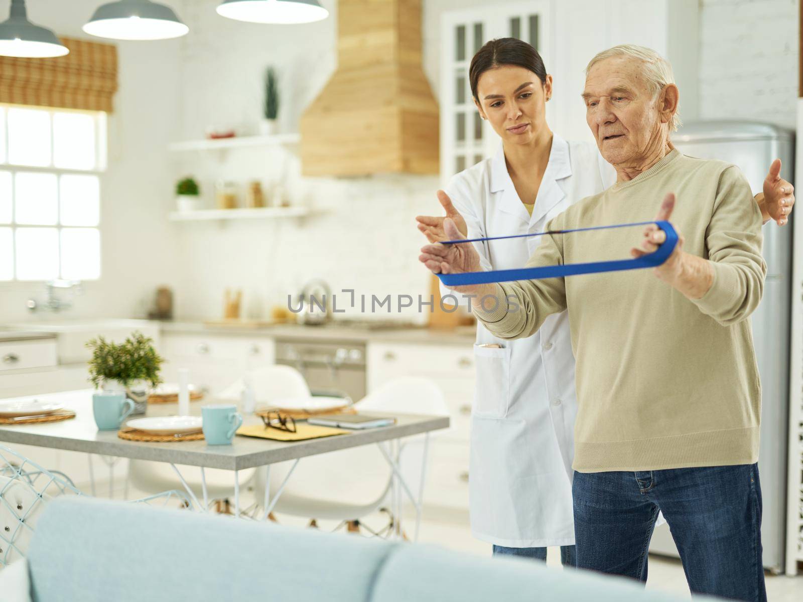 Young female doctor in a white coat explaining to a pensioner how to do exercises at home with fitness rubber