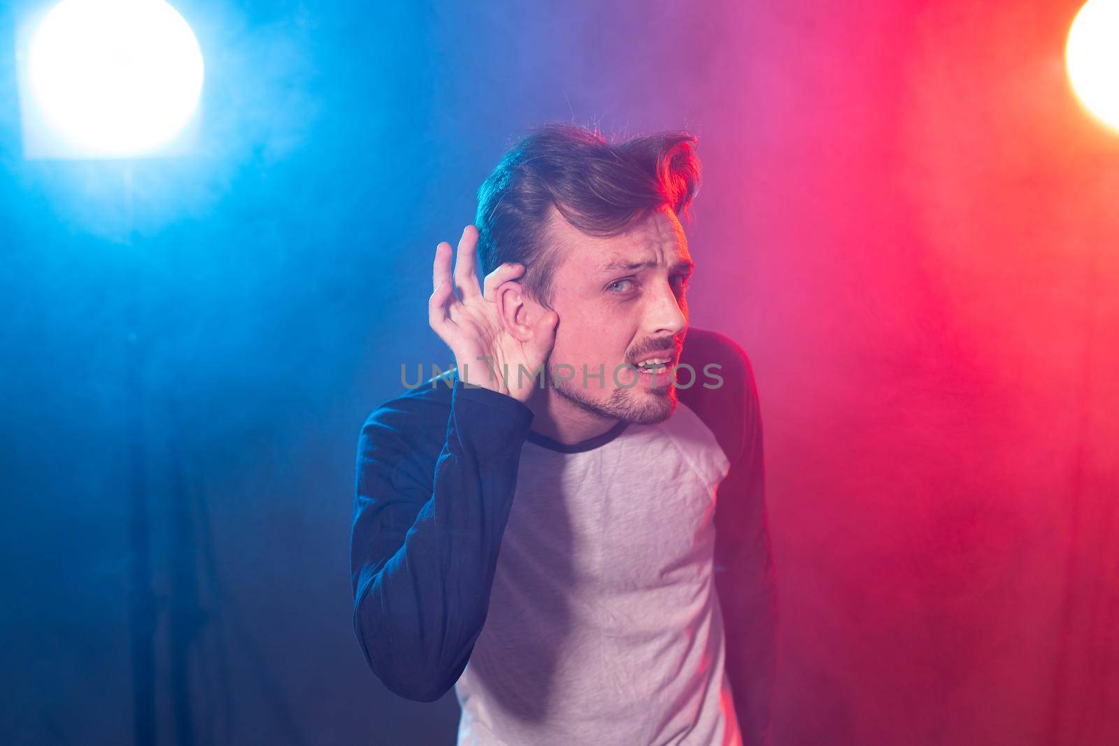 Young embarrassed handsome man hipster with a beard leaned his hand to his ear trying to listen to pose on a red blue background. Concept of importance of opinion