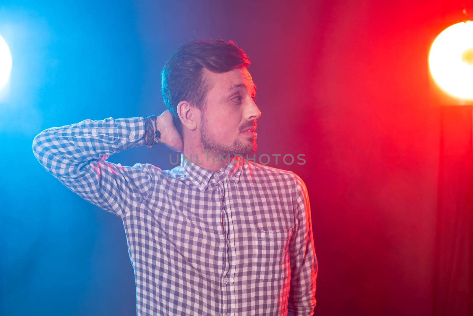 Carming young smiling hipster man is posing against the background of red and blue studio lighting. Concept of pensive man