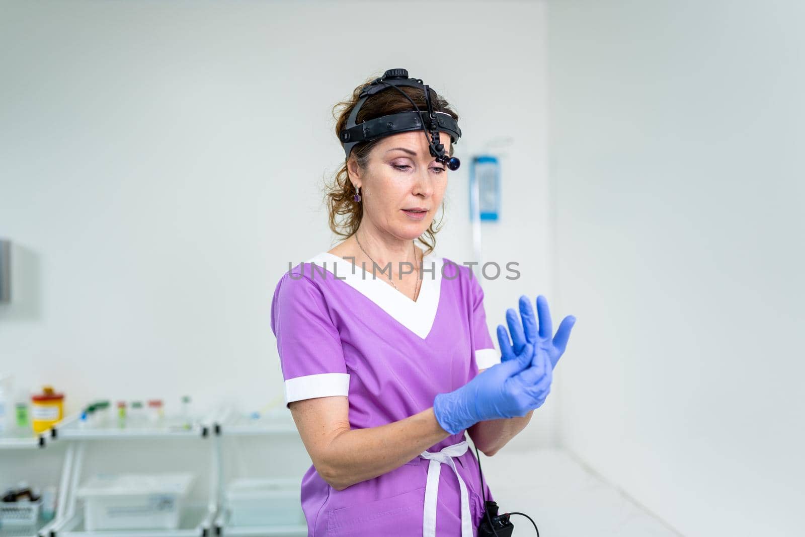 Portrait of confident female doctor. Female ENT doctor in purple medical clothes posing in office of the clinic on her head with a sedecin instrument to examine the patient. Healthcare occupations by Tomashevska