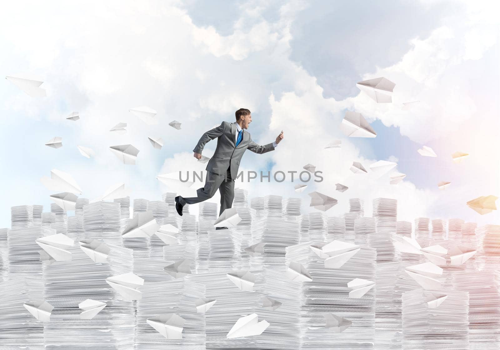 Businessman in black suit running with phone in hand among flying paper planes with cloudly skyscape on background. Mixed media.