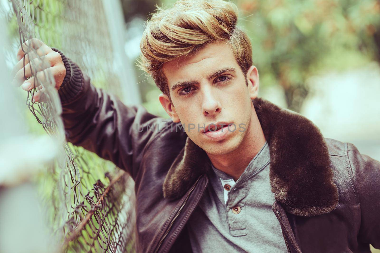 Portrait of a young handsome man, model of fashion, with modern hairstyle in urban background wearing aviator leather jacket. Blonde hair