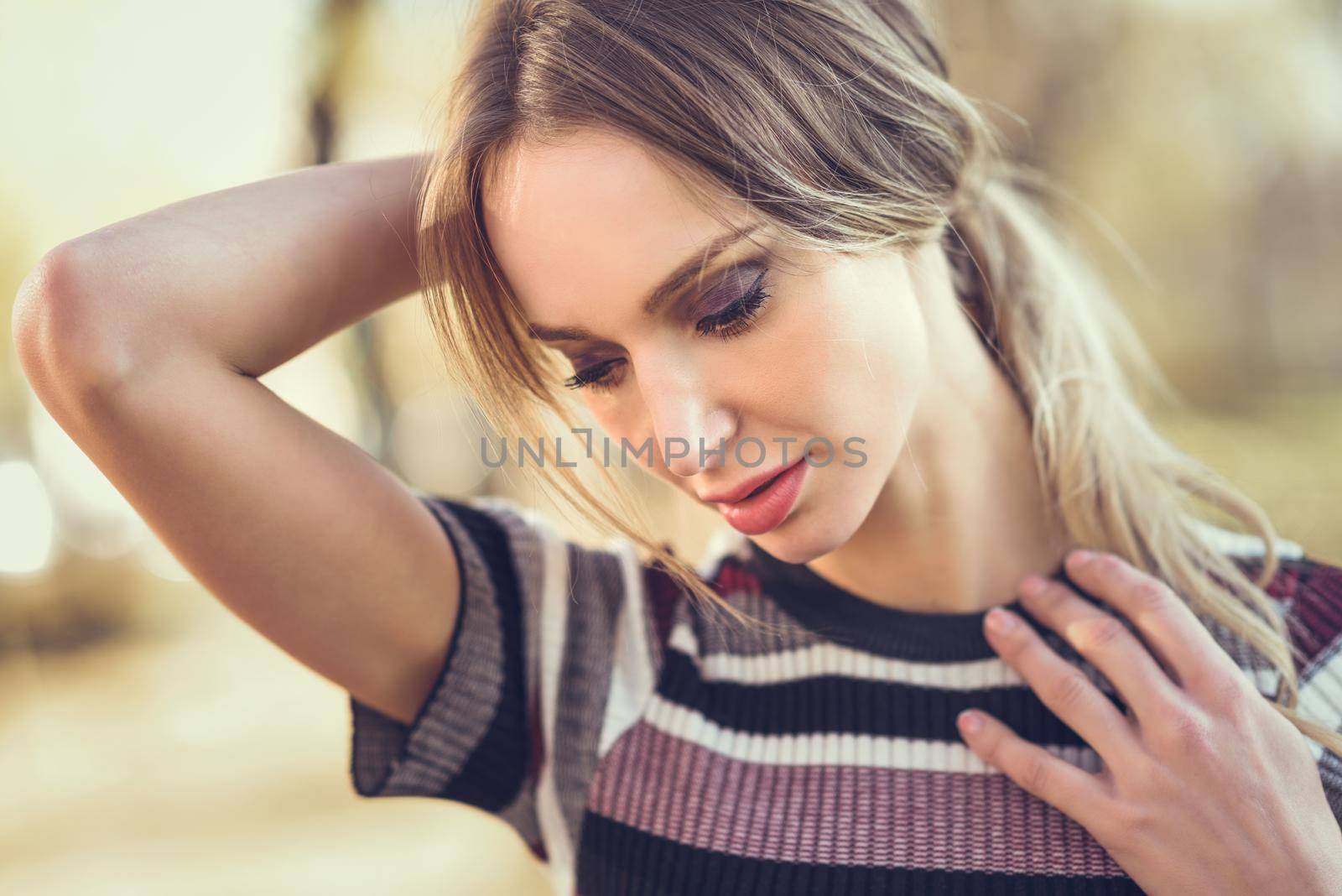 Close-up portrait of pensive young blonde woman standing in the street. Beautiful girl in urban background wearing striped sweater. Female with straight hair.