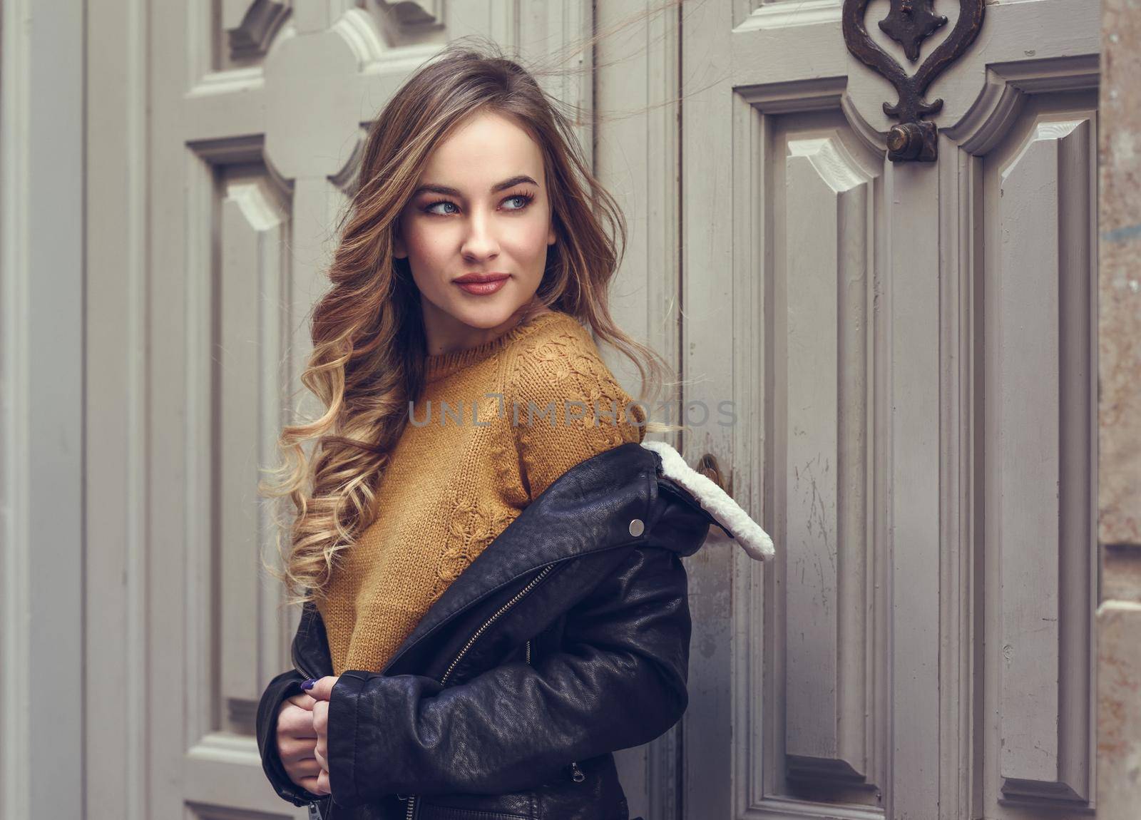 Blonde woman in urban background. Beautiful young girl wearing black leather jacket and mini skirt standing in the street. Pretty russian female with long wavy hair hairstyle and blue eyes.