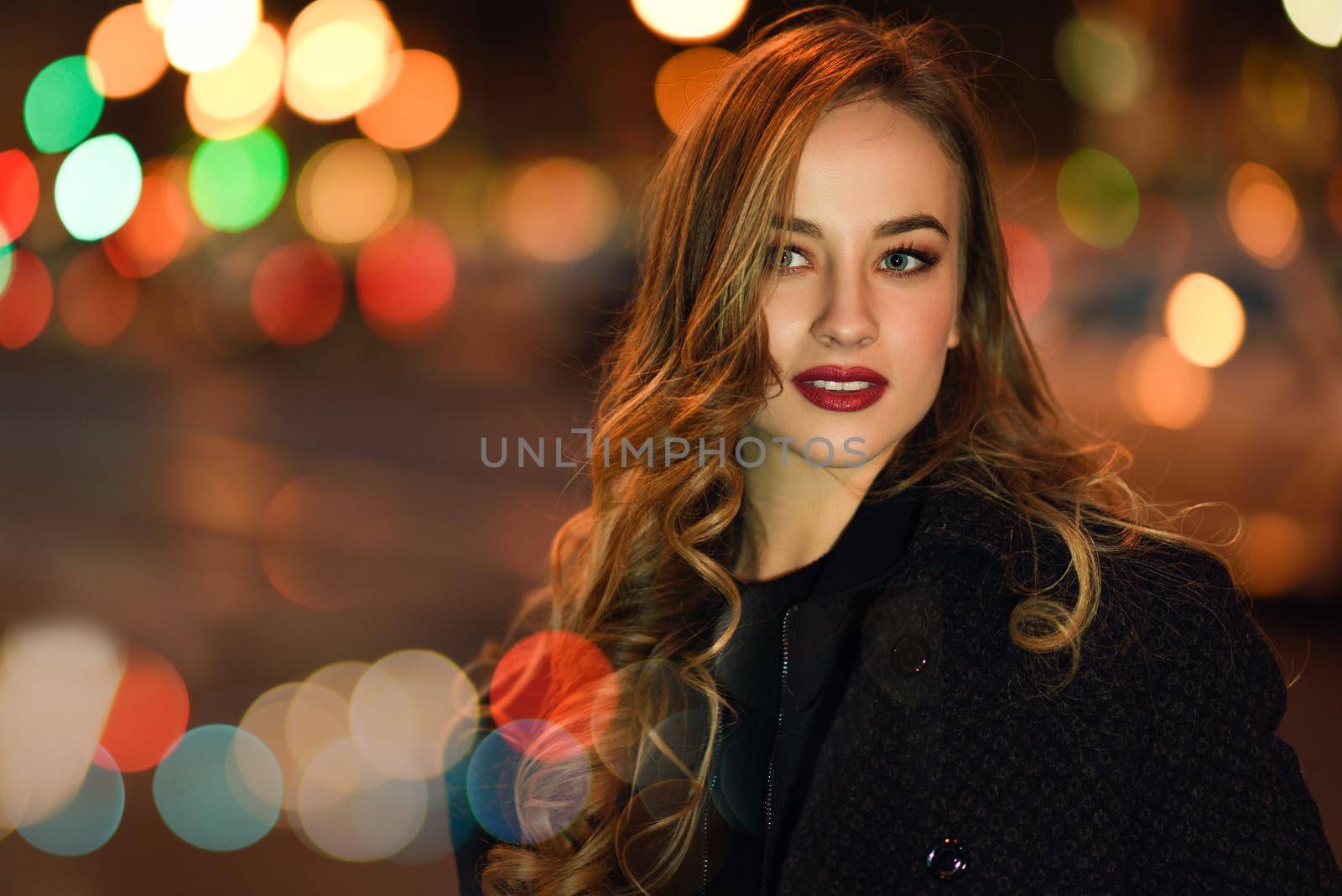 Beautiful young girl wearing black jacket walking in the street with defocused city lights at the background. Pretty russian female with long wavy hair hairstyle. Blonde woman in urban background.