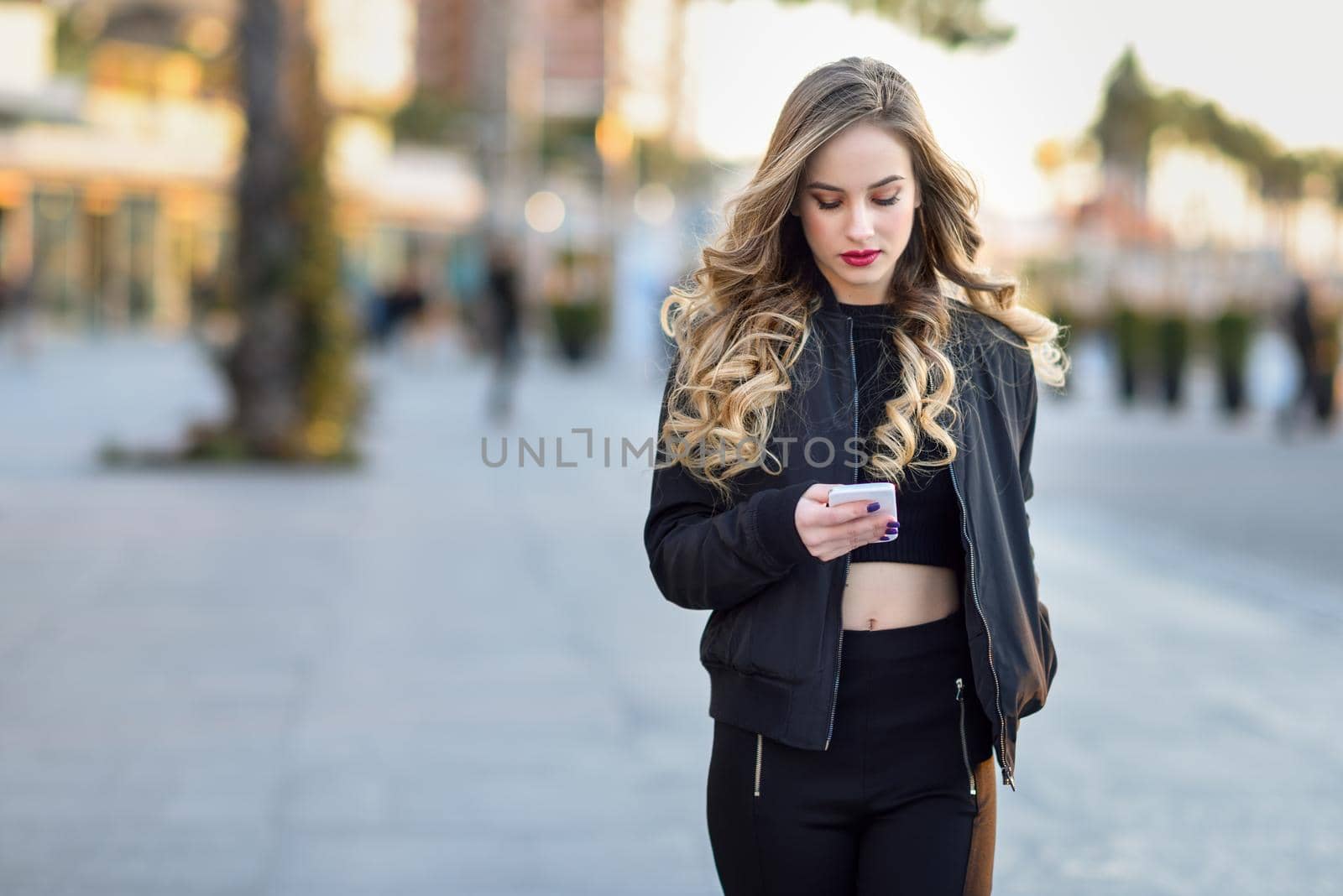 Blonde woman texting with her smart phone in urban background. Beautiful young girl wearing black jacket walking in the street. Pretty russian female with long wavy hair hairstyle.