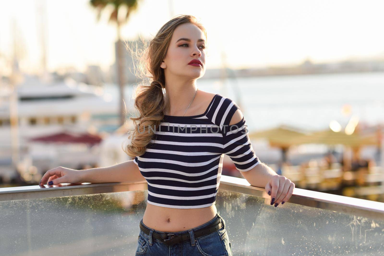 Blonde woman, model of fashion, standing in a harbor with boats and sea in the background. Beautiful young girl wearing striped t-shirt and blue jeans. Pretty russian female with pigtail.