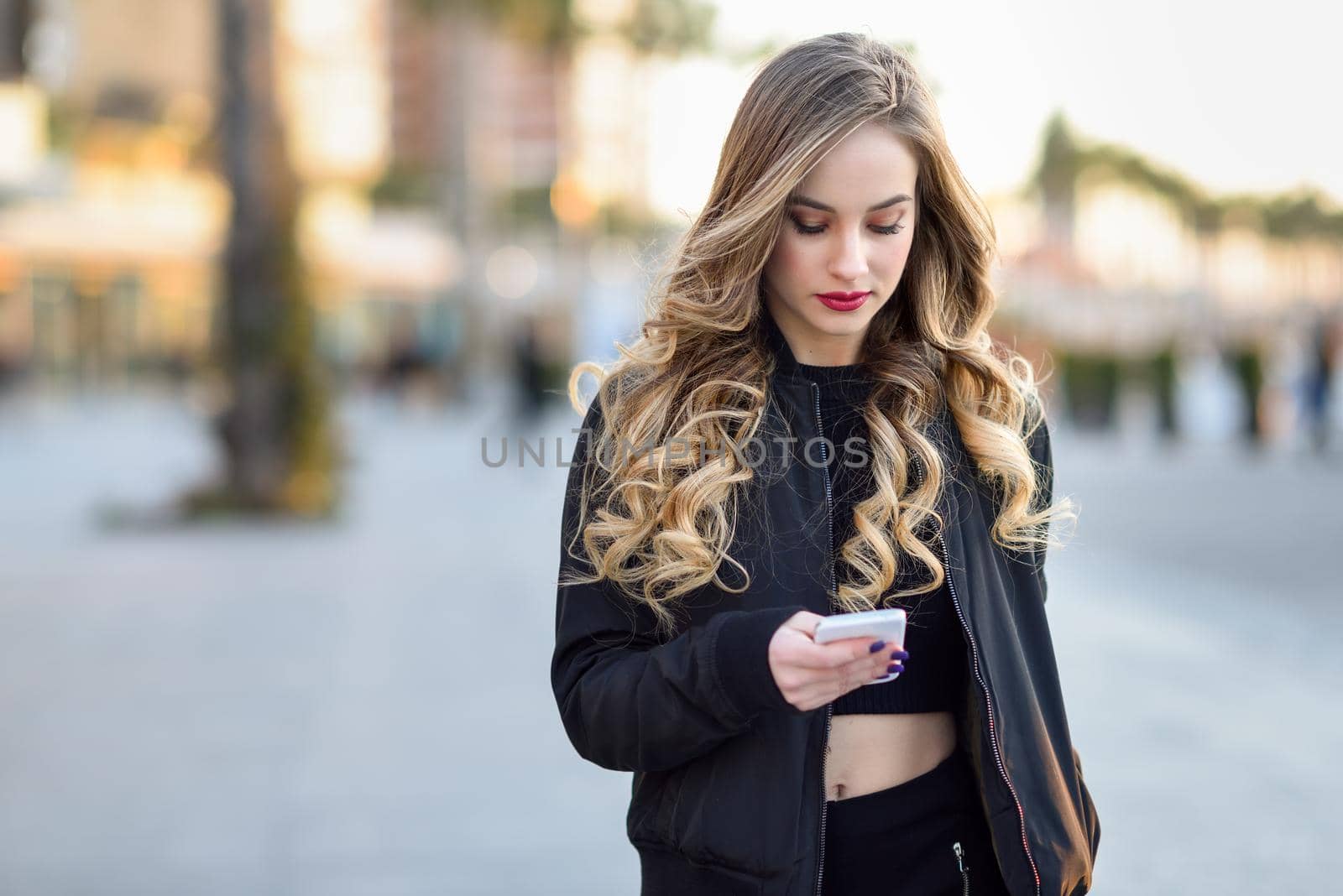 Blonde woman texting with her smart phone in urban background. Beautiful young girl wearing black jacket walking in the street. Pretty russian female with long wavy hair hairstyle.