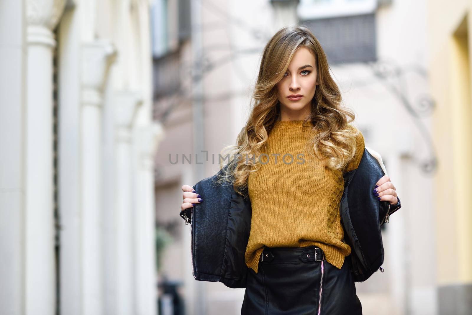 Blonde woman in urban background. Beautiful young girl wearing black leather jacket and mini skirt standing in the street. Pretty russian female with long wavy hair hairstyle and blue eyes.