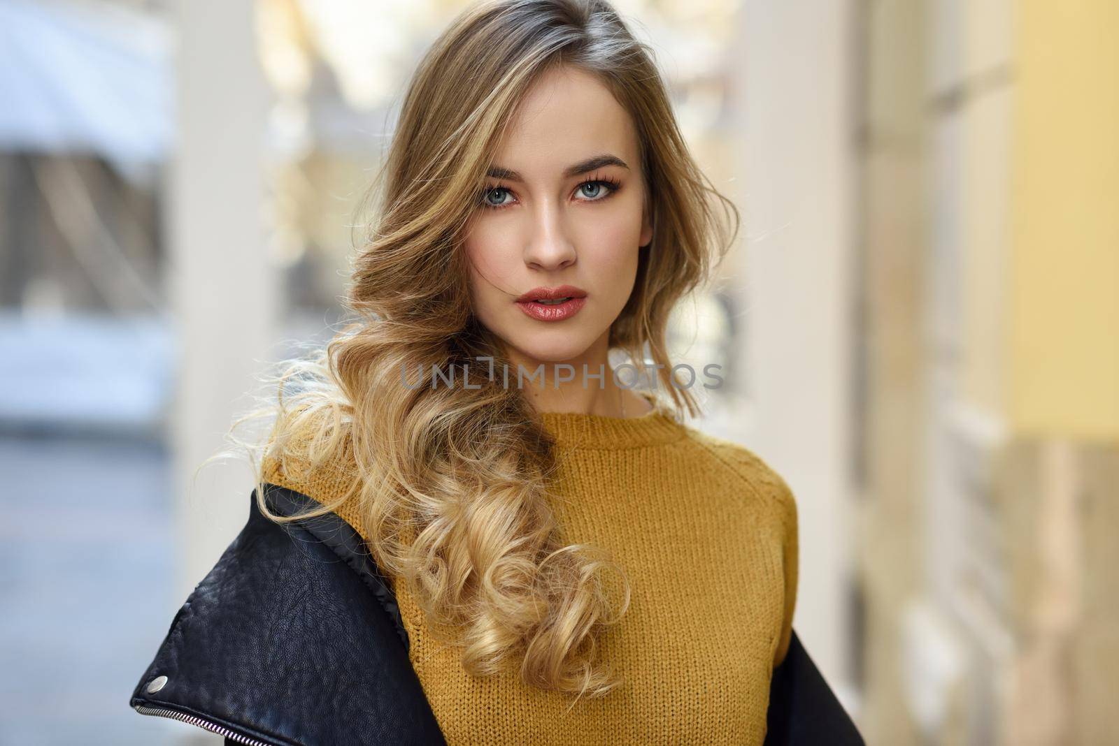 Blonde woman in urban background. Beautiful young girl wearing black leather jacket and mini skirt standing in the street. Pretty russian female with long wavy hair hairstyle and blue eyes.