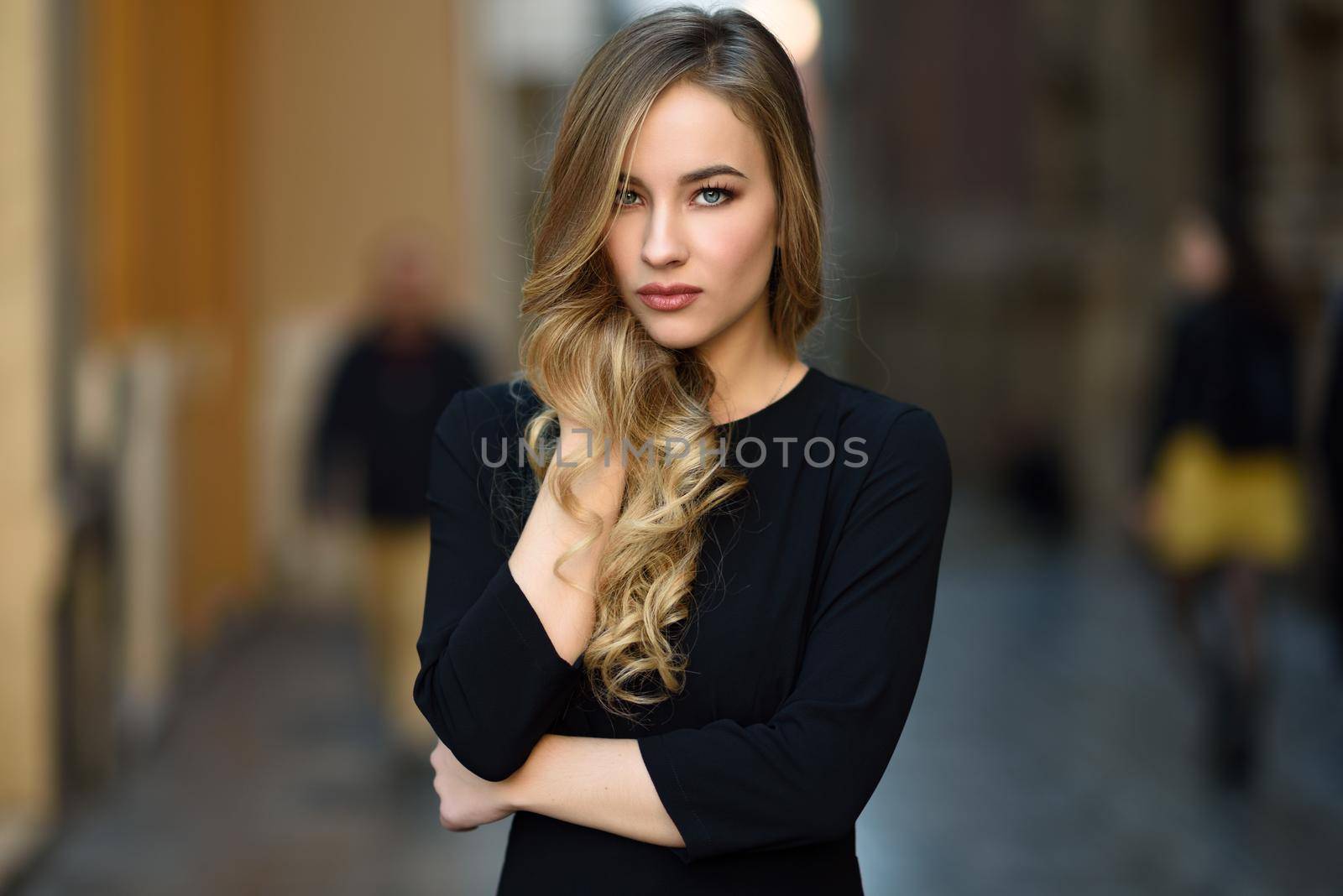 Blonde woman in urban background. Beautiful young girl wearing black elegant dress standing in the street. Pretty russian female with long wavy hair hairstyle and blue eyes.