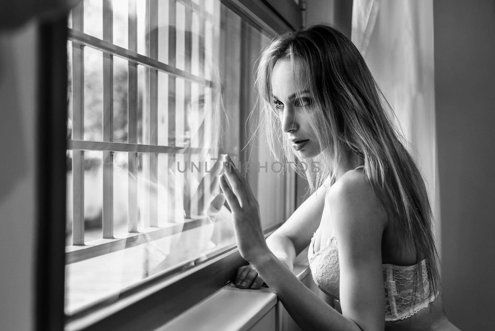 Beautiful blonde woman in white lingerie posing near a window. Young girl wearing underwear with long hair. Black and white photograph.