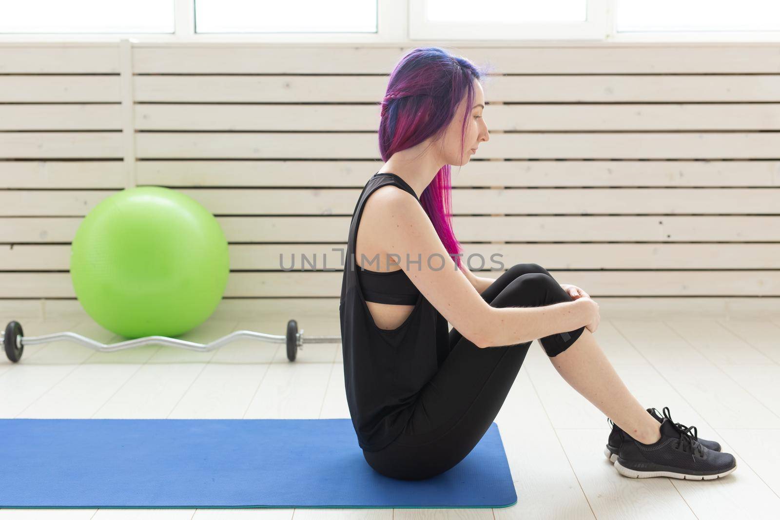 Young slim girl with colored hair makes an headstand on a sports blue mattress next to a barbell and green fitball. Concept of good physical fitness and regular training in the gym